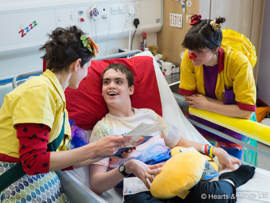 Two Clowndoctors wearing yellow coats stand on either side of a young patient who is smiling at the Clowndoctors from his hospital bed.